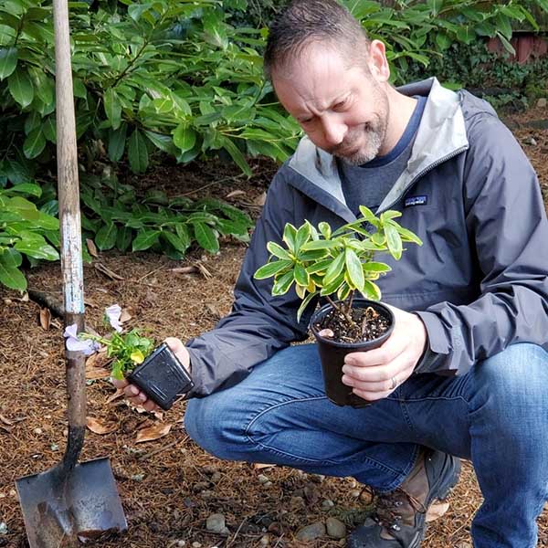 Man looking at plants in pots he is holding in his hands while squatting down towards the ground.